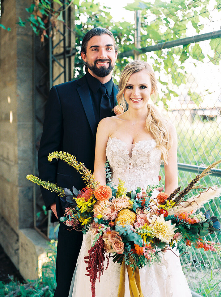 Bride and groom, wearing a black tuxedo and white wedding gown, smile with a large bouquet of yellow, blush and orange flowers.