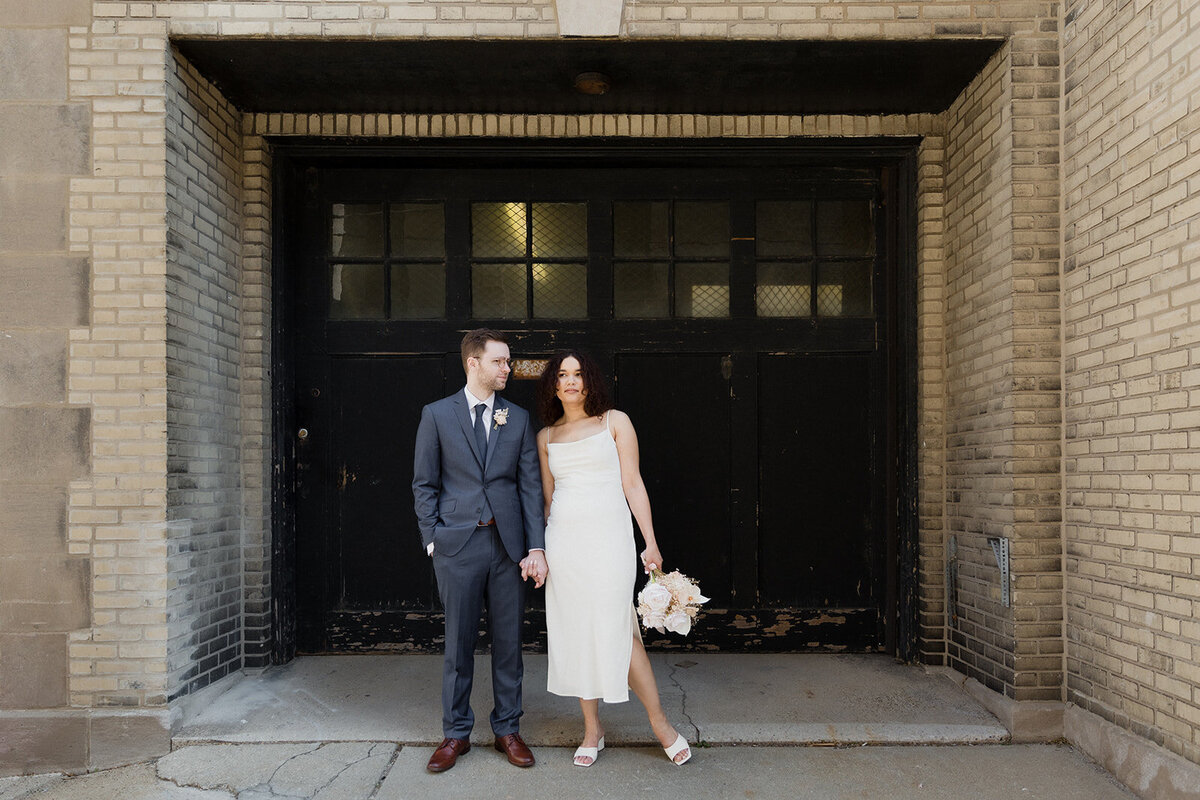 Just Married photo session couple stands in front of a black garage door and brick wall and hold hands, groom is looking at bride as she looks on.