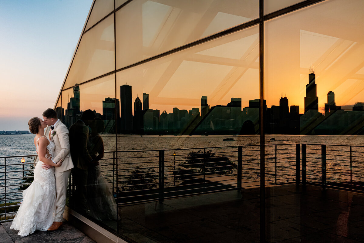 Bride and groom kiss with the chicago skyline reflected in the window at sunset at  the adler planetarium in Chicago, Illinois
