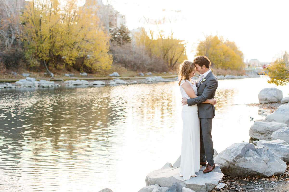 Couple embracing on the rocks near the river at River Cafe, a riverside wedding venue in downtown Calgary, featured on the Brontë Bride Vendor Guide.