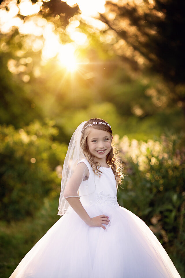 A young girl stands proudly with a hand on her hip in a large white communion dress in a park at sunset