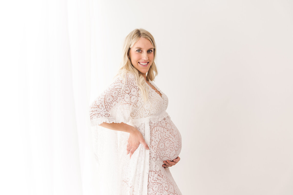 A joyful pregnant woman in a lace dress lovingly cradling her belly with a bright smile, against a clean, white backdrop.