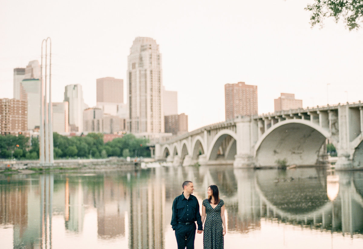 fiances hold hands over mississippi river with minneapolis skyline in the background