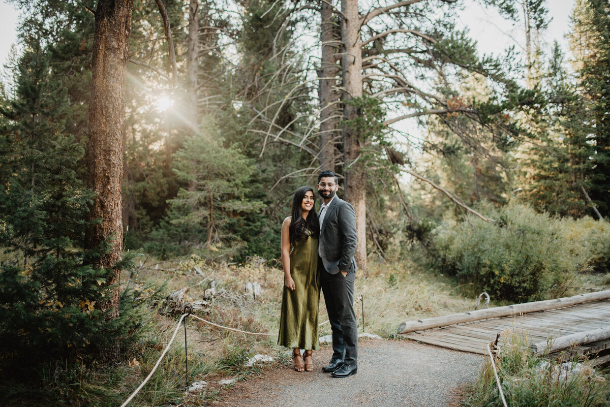 photographers in jackson hole photographs engagement session in Jackson Hole Wyoming  with the sunshine gleaming through the trees as the couple stands on a wooden trail