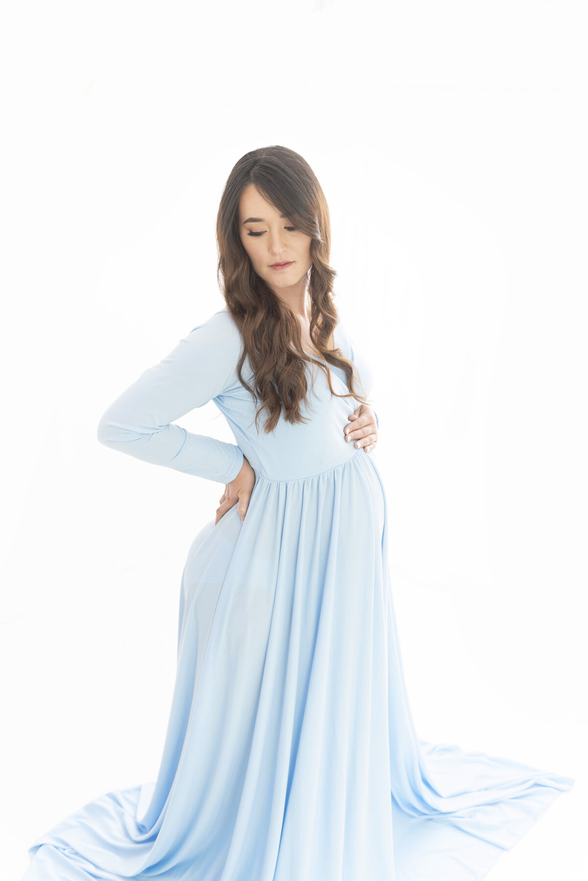 A pregnant woman stands in a studio gazing down her shoulder in a blue maternity gown