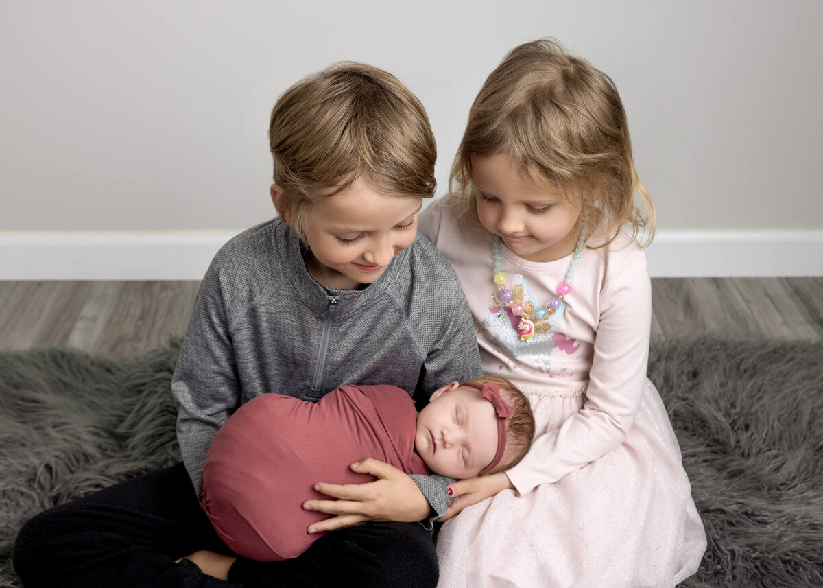 brother and sister holding their newborn baby sister and looking down at her while sitting on a grey shaggy rug