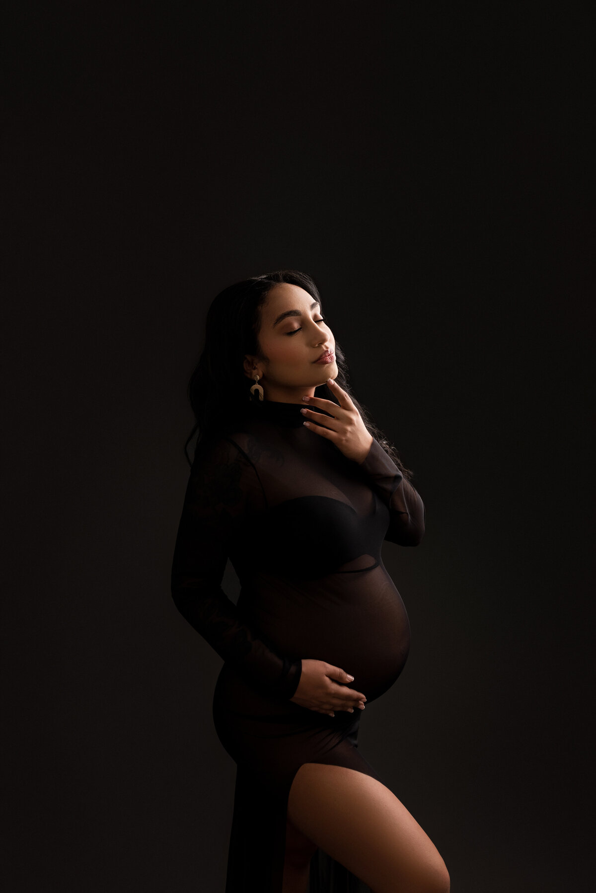 Maternity photo captured by Katie Marshall, a leading maternity photographer in New Jersey. The image features a woman standing at an angle away from the camera, adorned in a long black body-con maternity dress with a stylish slit. Her foreleg is gracefully bent, and her forearm supports her baby bump. Her back arm gently touches her chin. With closed eyes and her head tilted towards the light source, dark shadows artfully create drama and depth, resulting in a striking and emotive maternity composition.