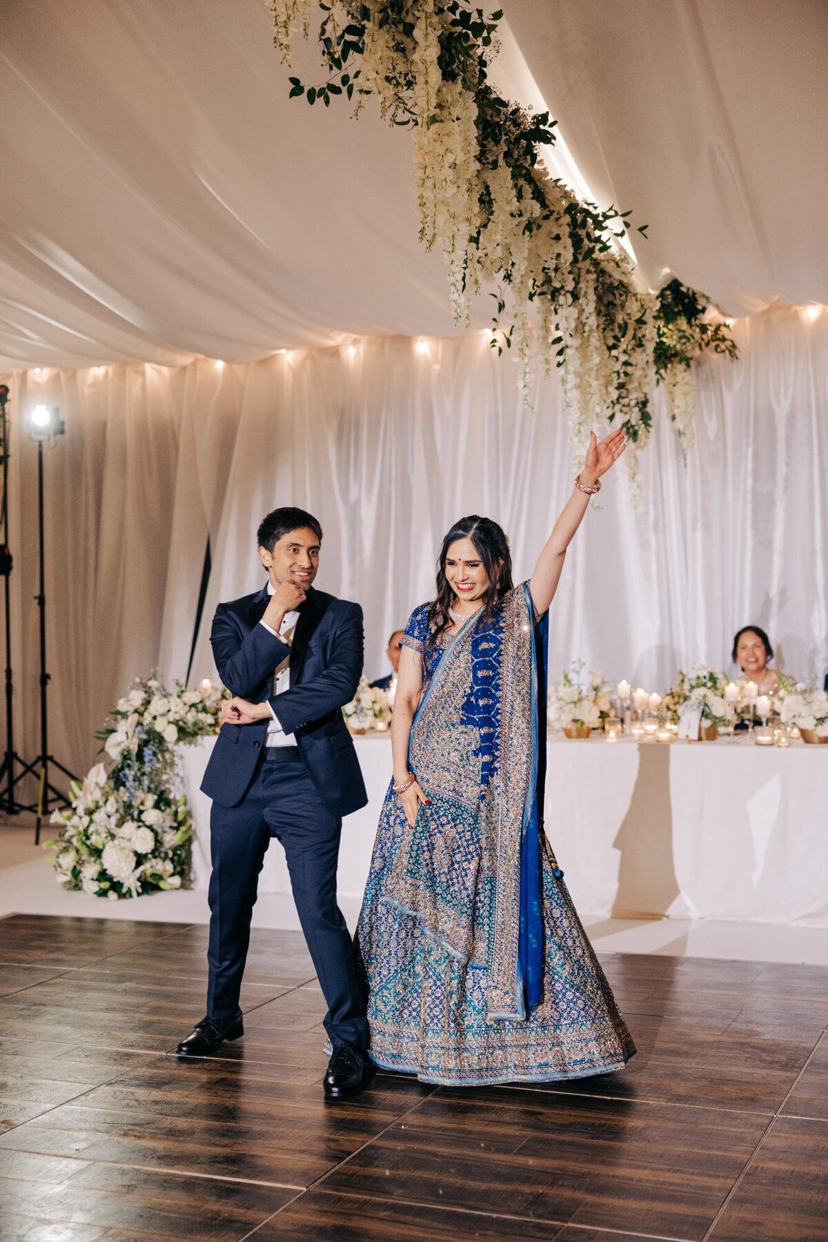 A couple dancing energetically at their wedding reception, with the bride in a decorative blue sari and the groom in a suit, under a canopy adorned with flowers.