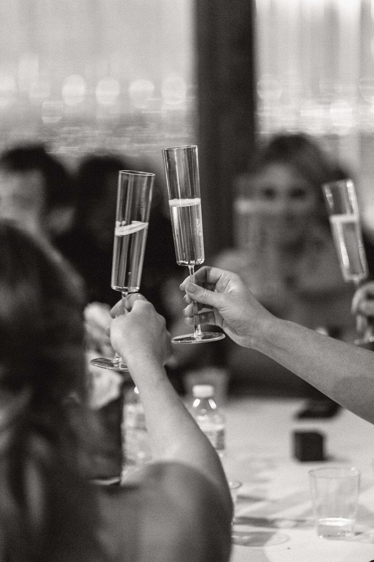 A black and white photograph of guests during the reception on Lindsay and Tom’s wedding day at Thompson Island in Boston, Massachusetts. The photo is focused on the champagne flutes as guests cheers each other during the toasts. Wedding photography by Stacie McChesney/Vitae Weddings.
