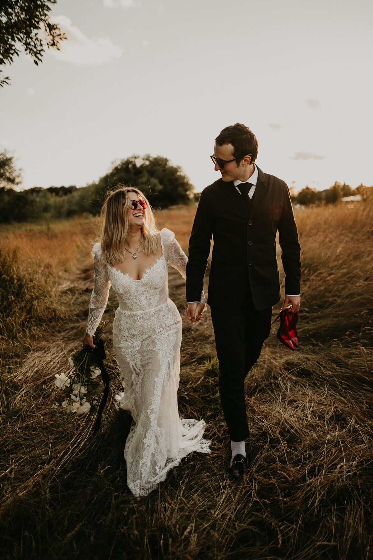 A groom carries his brides red christian louboutin heels as they walk through the long grass of a farm wedding venue.