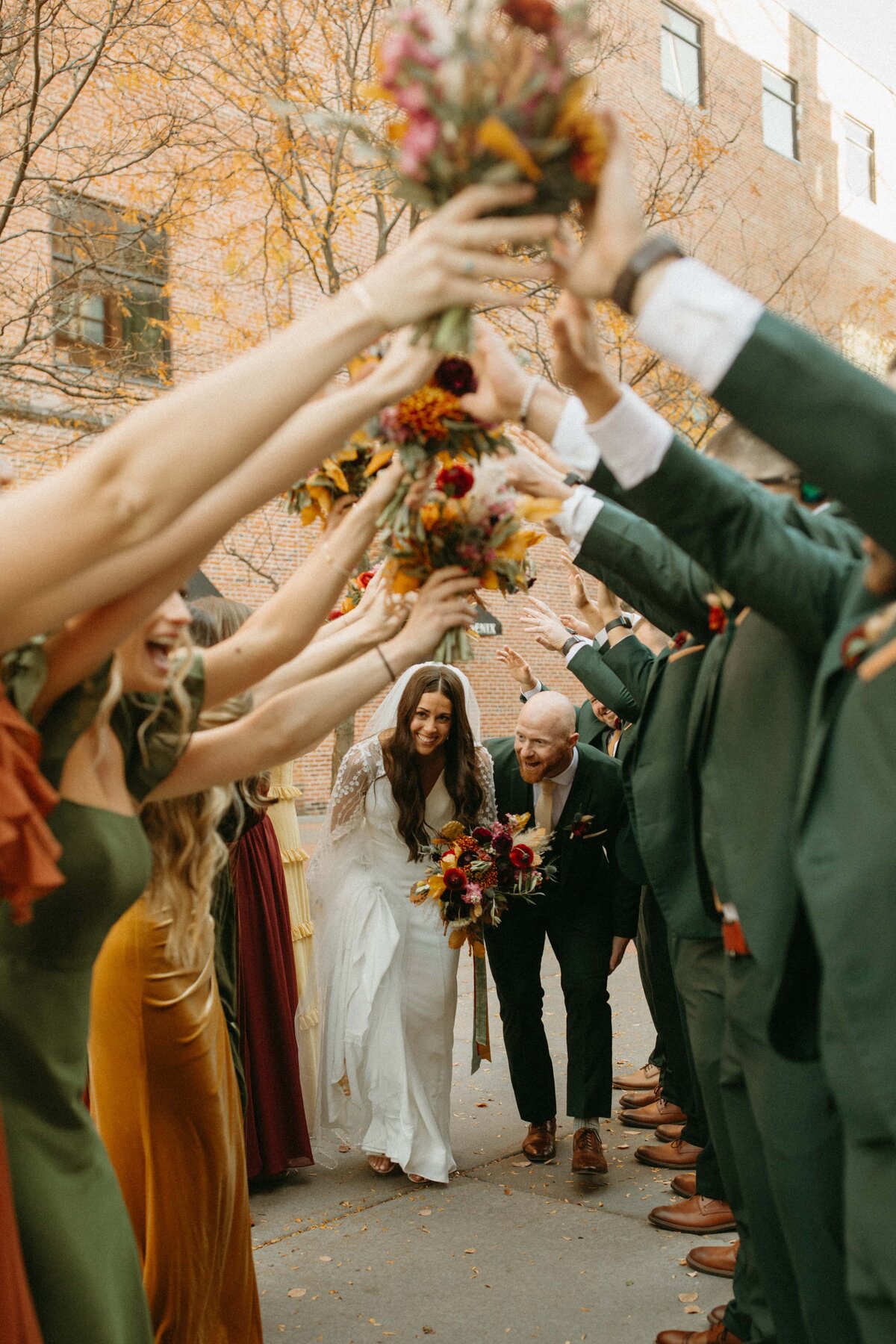 A bride and groom walk through a cheering crowd of guests at a park farm winery wedding, holding up flower bouquets, creating a tunnel of floral arches.