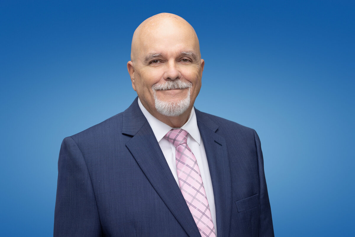 An older bald man with a beard wears a blue suit and pink tie as he poses for a professional headshot picture on a blue background for Janel Lee Photography studios in downtown Cincinnati Ohio
