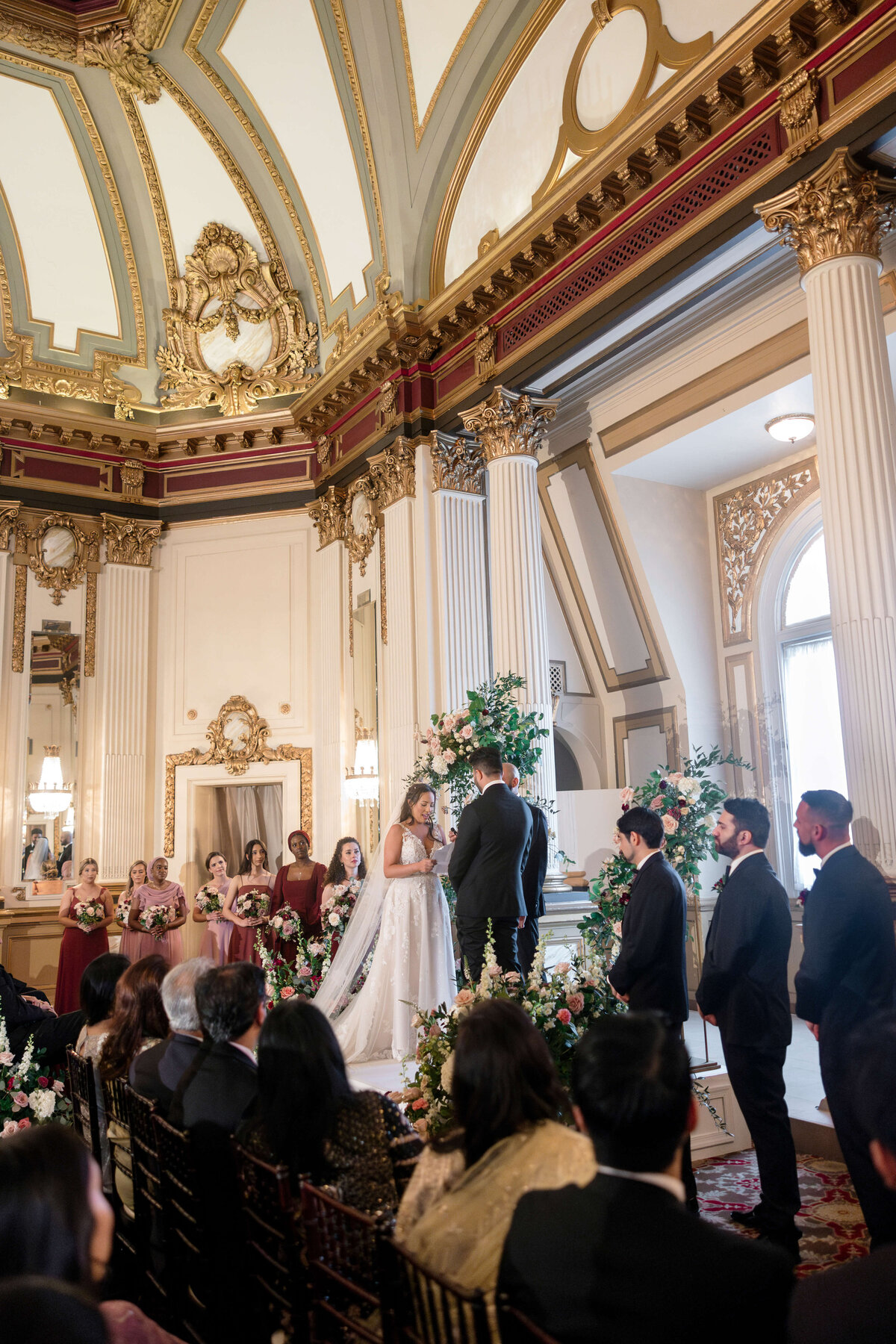 A bride and groom stand at the altar in an opulent wedding venue with ornate gold and white architecture. Bridesmaids in pink dresses and groomsmen in dark suits look on as guests watch from seated rows. Floral arrangements adorn the scene.