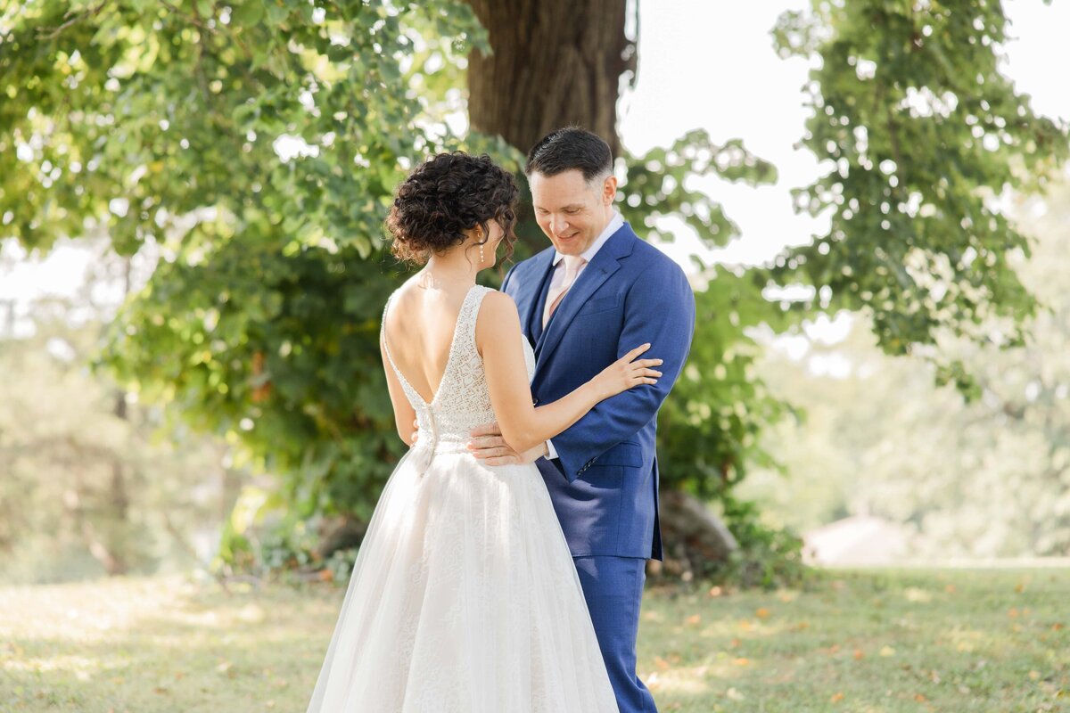 A bride and groom smiling at each other, holding hands in a sunlit park, with trees in the background during their park wedding.