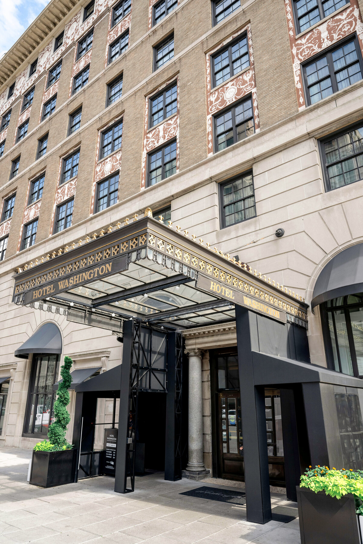 Entrance of the Hotel Washington with a grand black and gold marquee, framed by a classic stone building facade with large windows. Planters with greenery are positioned on each side of the entrance.