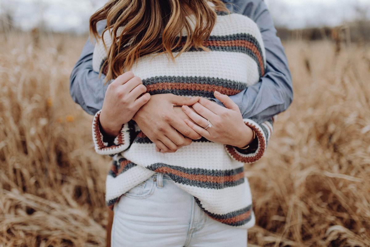 Close up on engaged couples arms and hands. Boy is behind girl, wrapping his arms around her, and girl has her hands on his arms, showing her engagement ring.