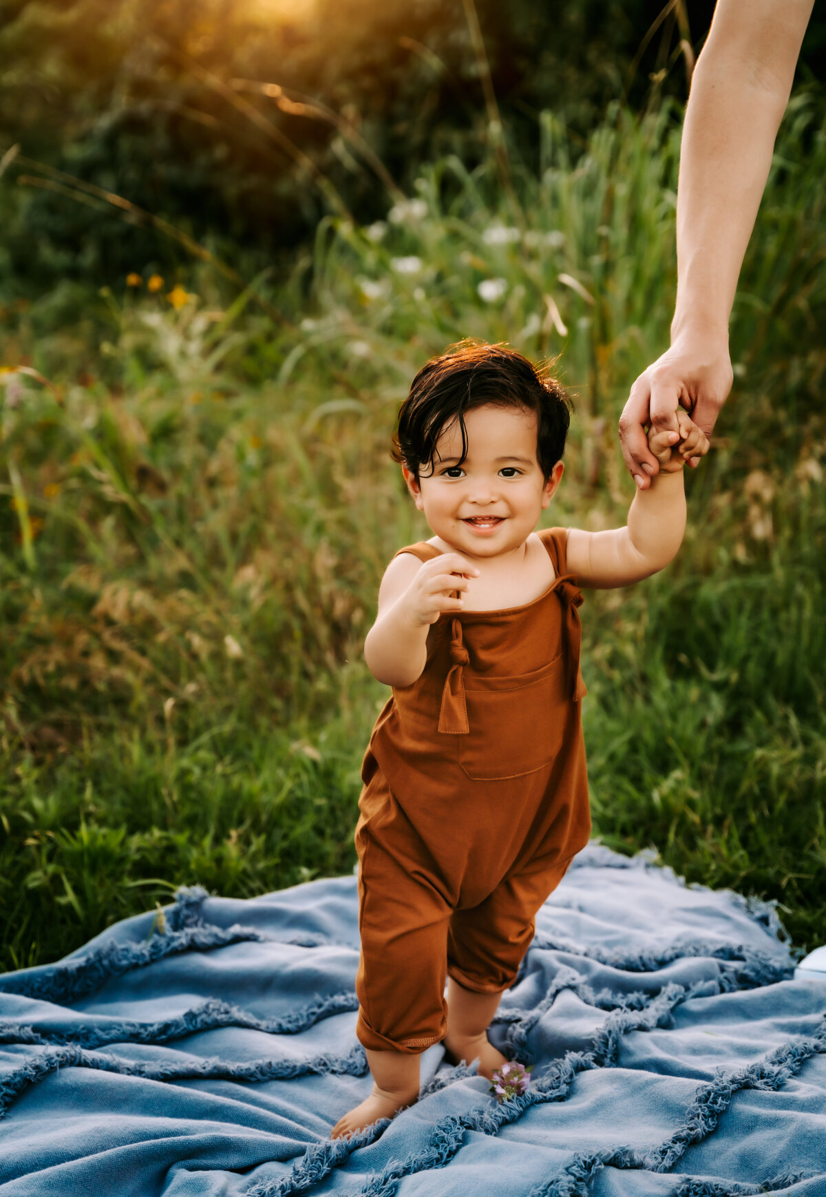 family photographer, a baby boy smiles standing on a picnic blanket with dad's help