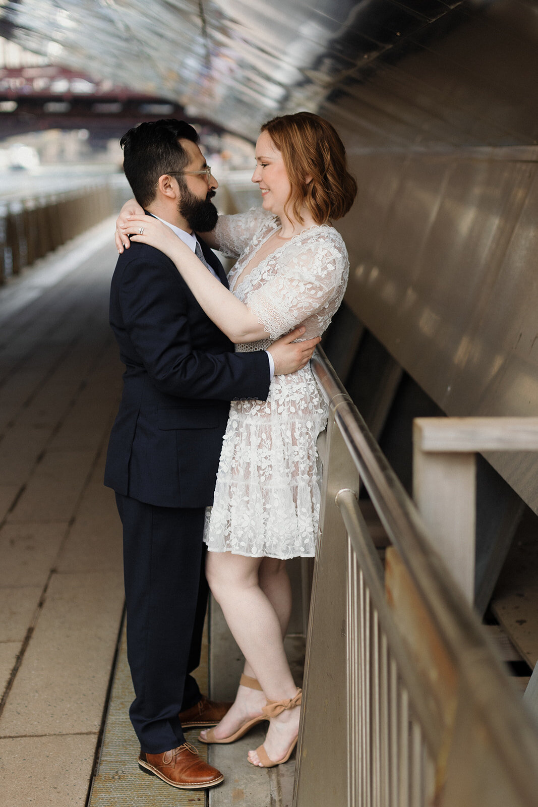 Wedding couple embraces while standing under Chicago River bridge. Bride leans against a rail while groom embraces her.