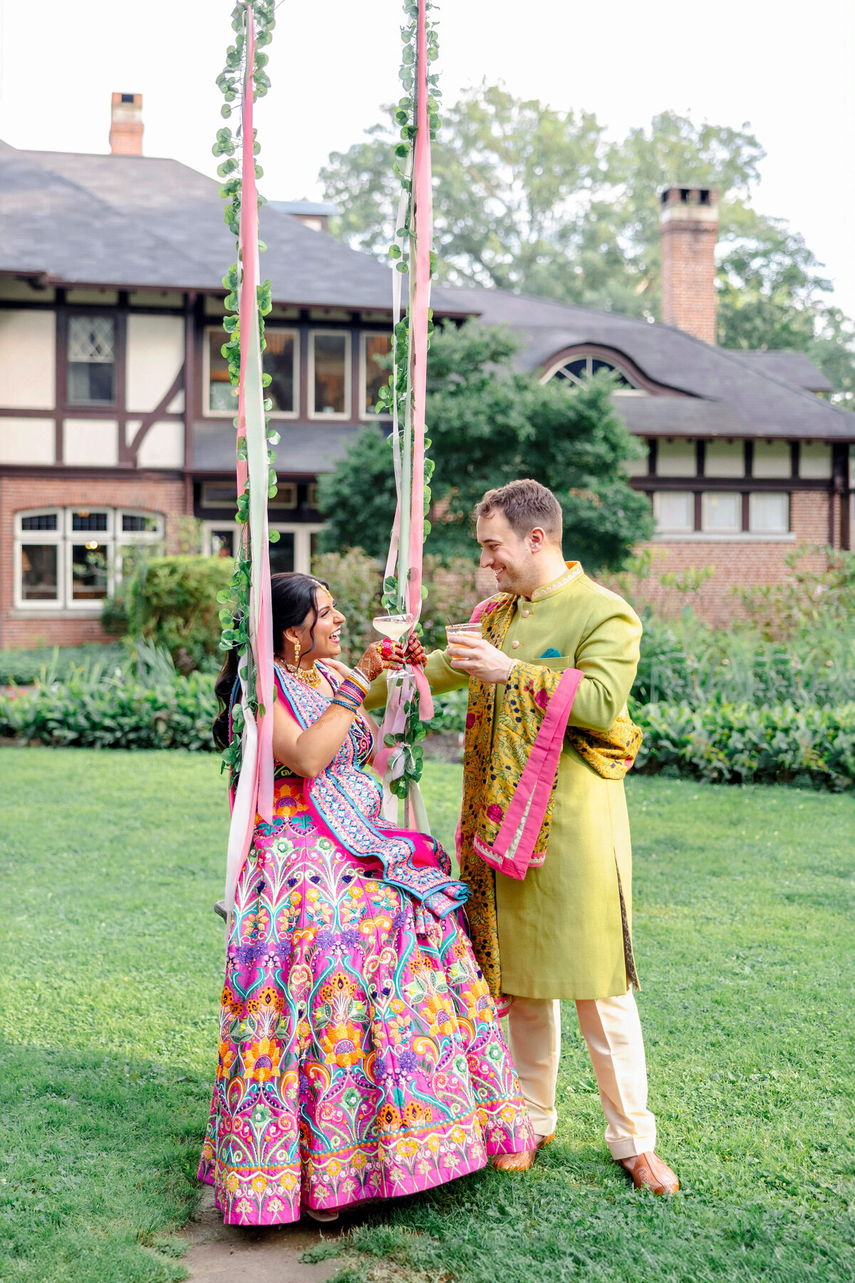 A couple in colorful traditional attire stands on a garden swing adorned with pink ribbons and flowers. They smile at each other, surrounded by greenery, in front of a charming Tudor-style house.