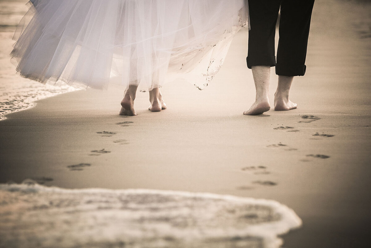 A bride and groom walk along the beach leaving footprints in the sand