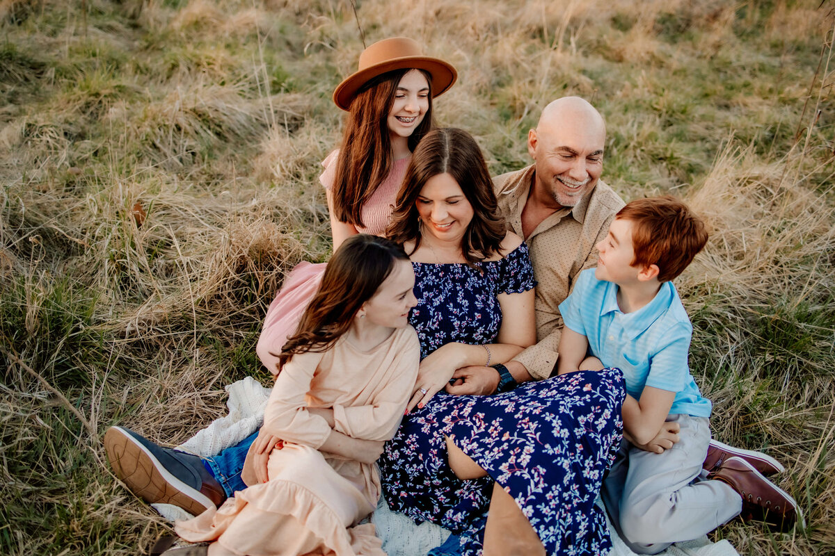 family of 5 snuggled on a blanket close together looking at each other in a field at sunset