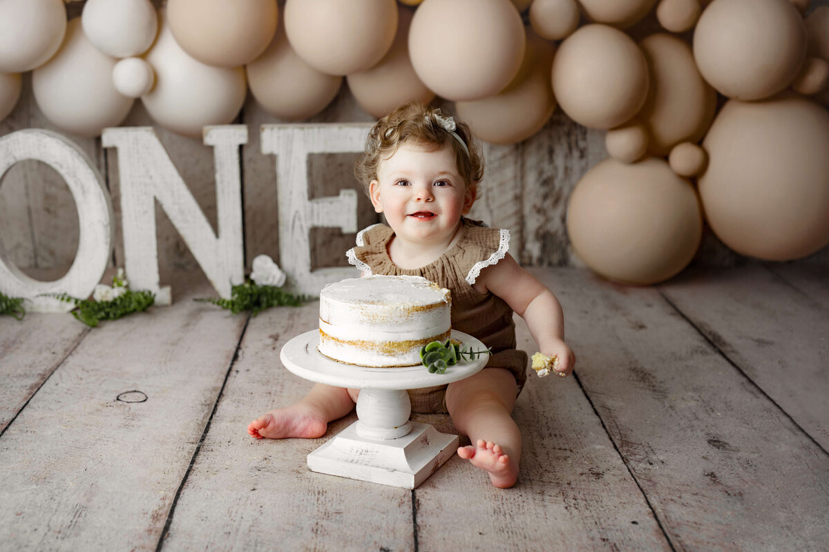 one year old girl sitting in front of her birthday cake in from of O-N-E letters and balloons