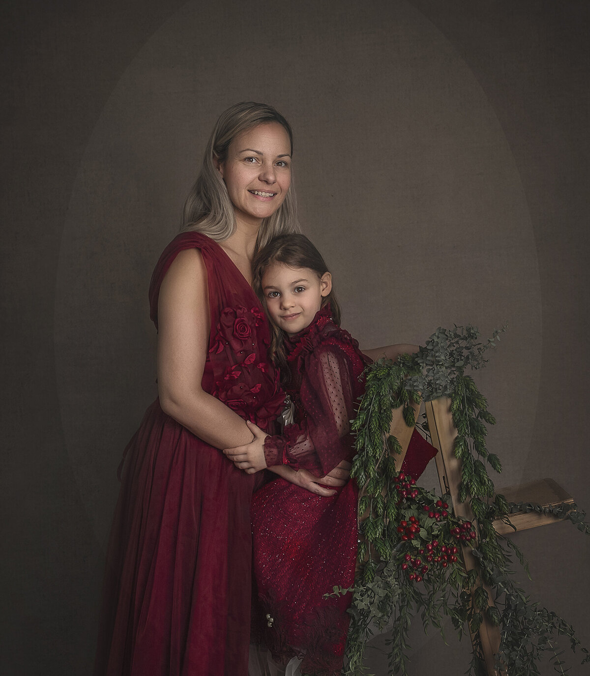 A mom and daughter both dressed in red dresses are posed on a ladder that has been decorated with green plants. Photo take in Ottawa.