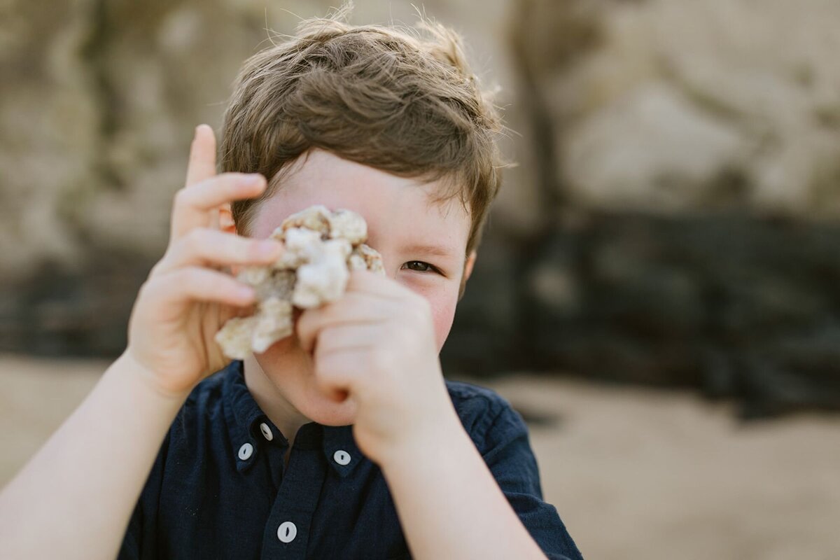 A little boy holds up a piece of coral to his eye playing on the beach.