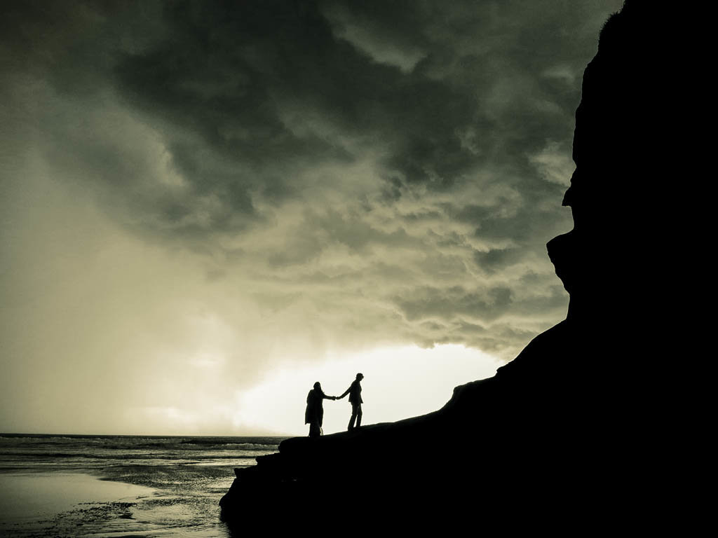 California coast adventure elopement  photography session.  Couple walking on rocks edge holding hands silhouetted with stormy sky above.