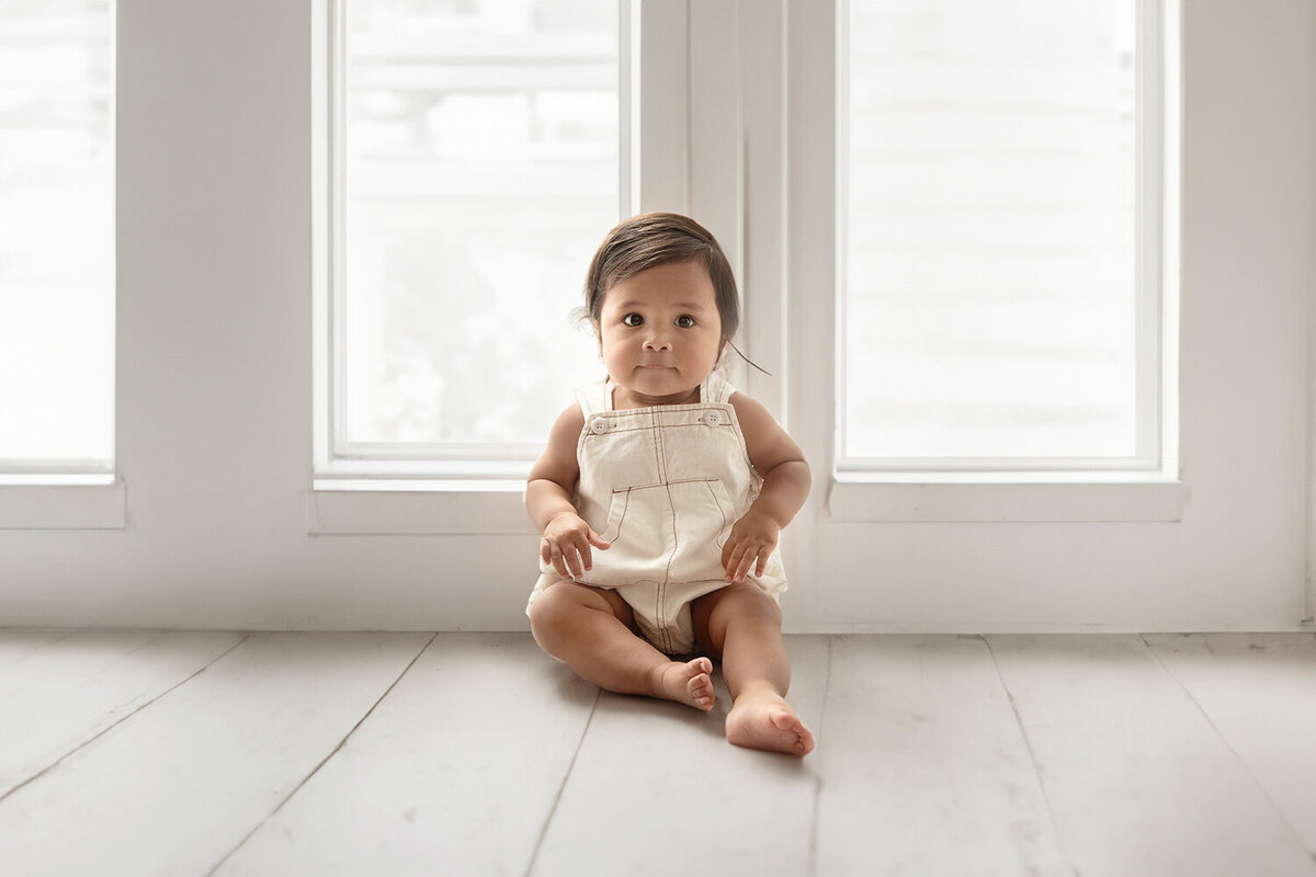 A young toddler in white overalls sits under some floor to ceiling windows in a studio