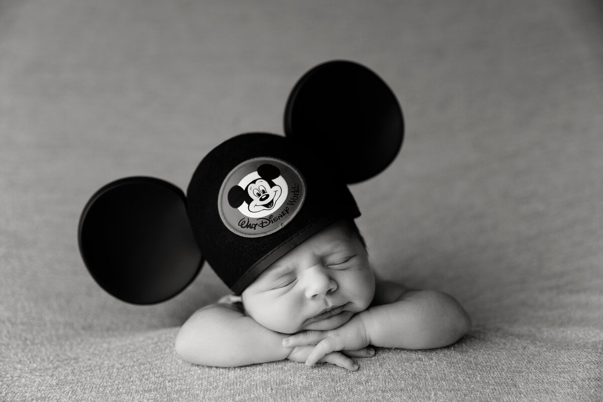A newborn baby sleeps on its hands wearing a Mickey Mouse ears hat