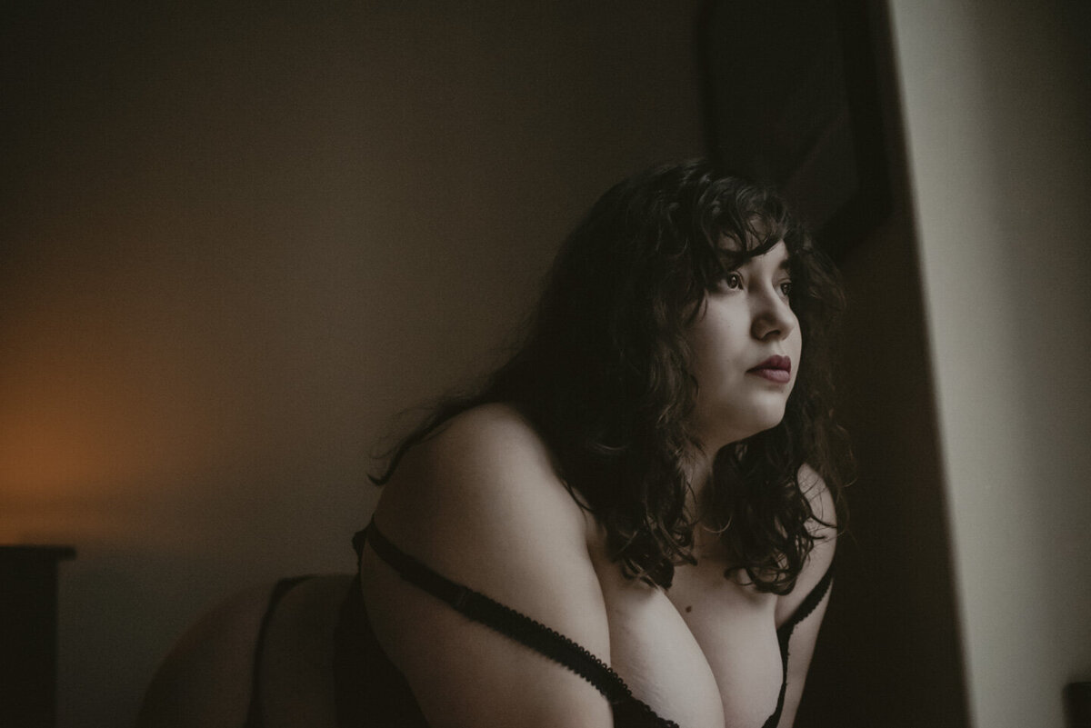 taeya in lingerie with her bra straps halfway down her arms. She is leaning on a chair, looking out her window. There is candle light adding warmth, in the left side of the frame, behind her.