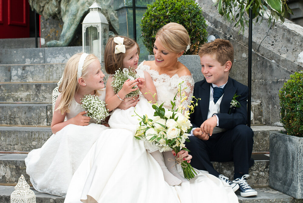 blonde bride wearing a scalloped lace, trumpet style wedding dress and white bouquet, sitting with her flower girls and page boy wearing black Converse trainers