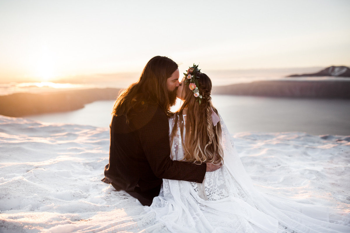 Couple in wedding attire sitting in the snow  and kissing a top an overlook.