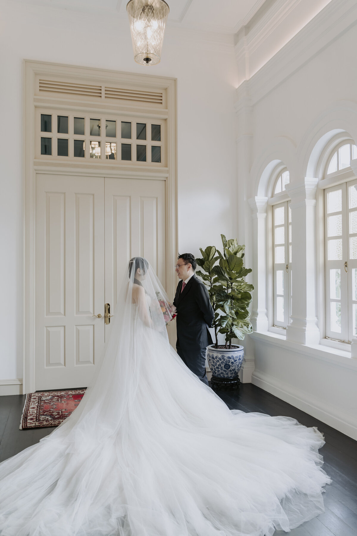 bride in white gown holding flowers and groom in a black suit stand in front of the window in the Raffles hotel