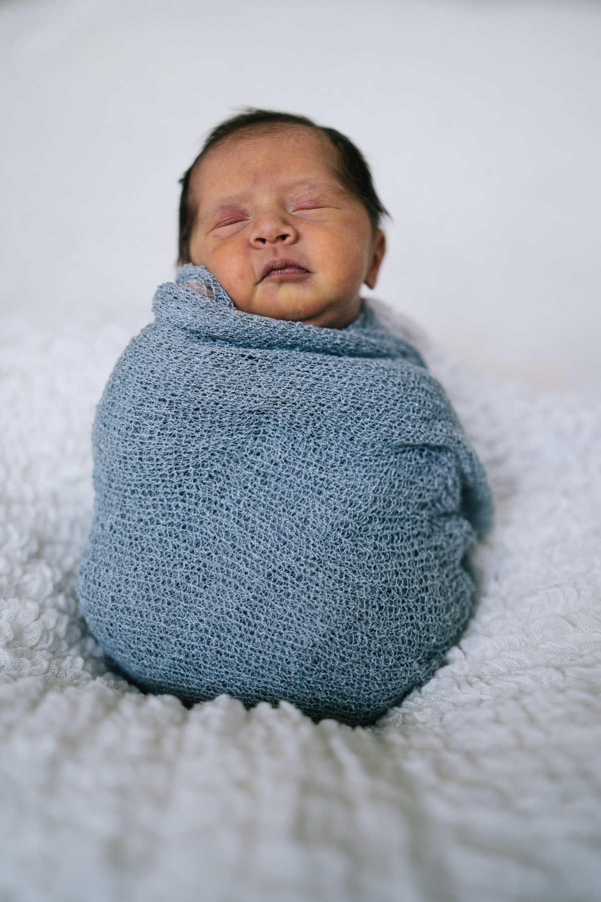 Newborn baby wrapped in cheesecloth blanket sitting on white blanket in San Antonio Photography studio.