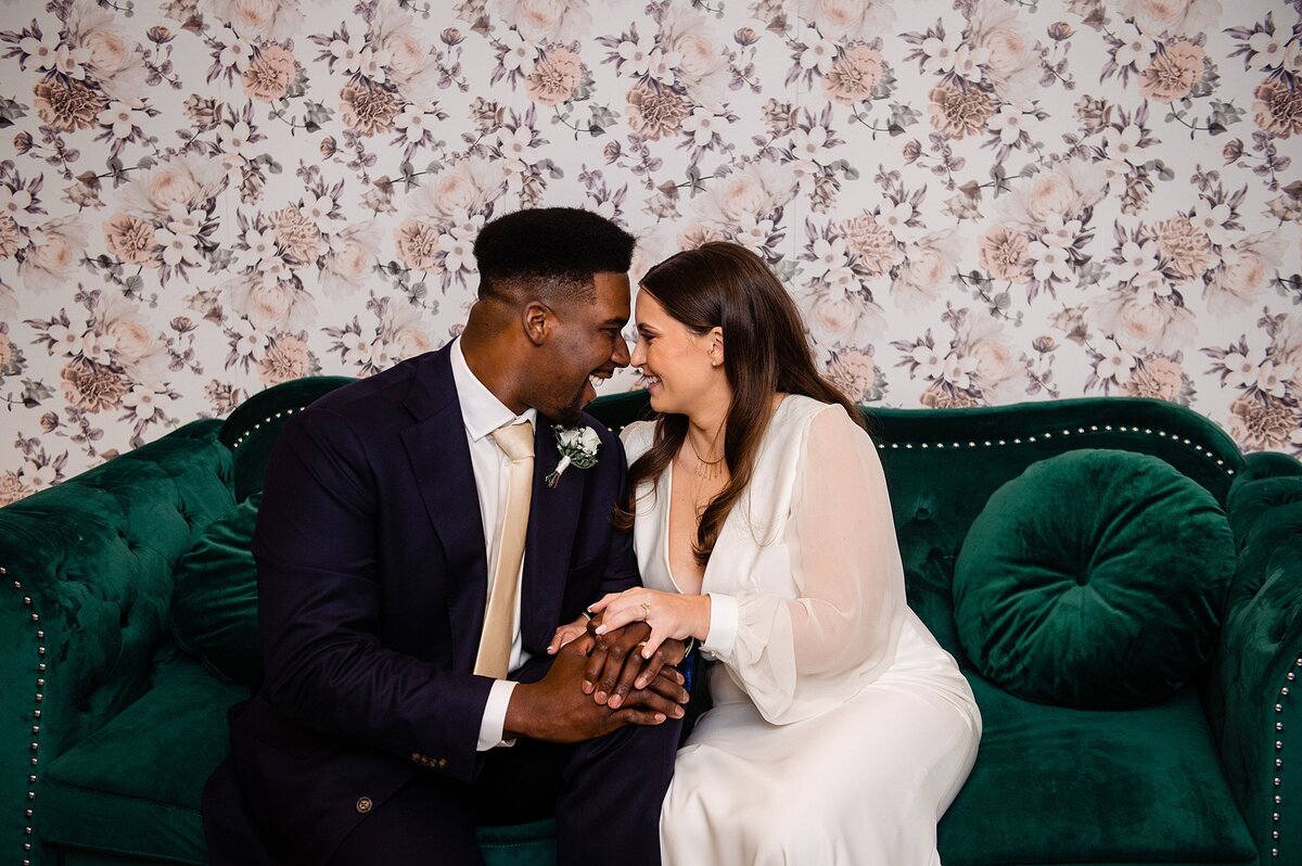 The African American groom wearing a black suti with a white shirt and blush tie sits on an emerald green velvet sofa holding hands with the bride wearing a long sleeved wedding dress with a plunging neckline. The wall behind them has blush colored floral wall paper at Steel Magnolia Barn.