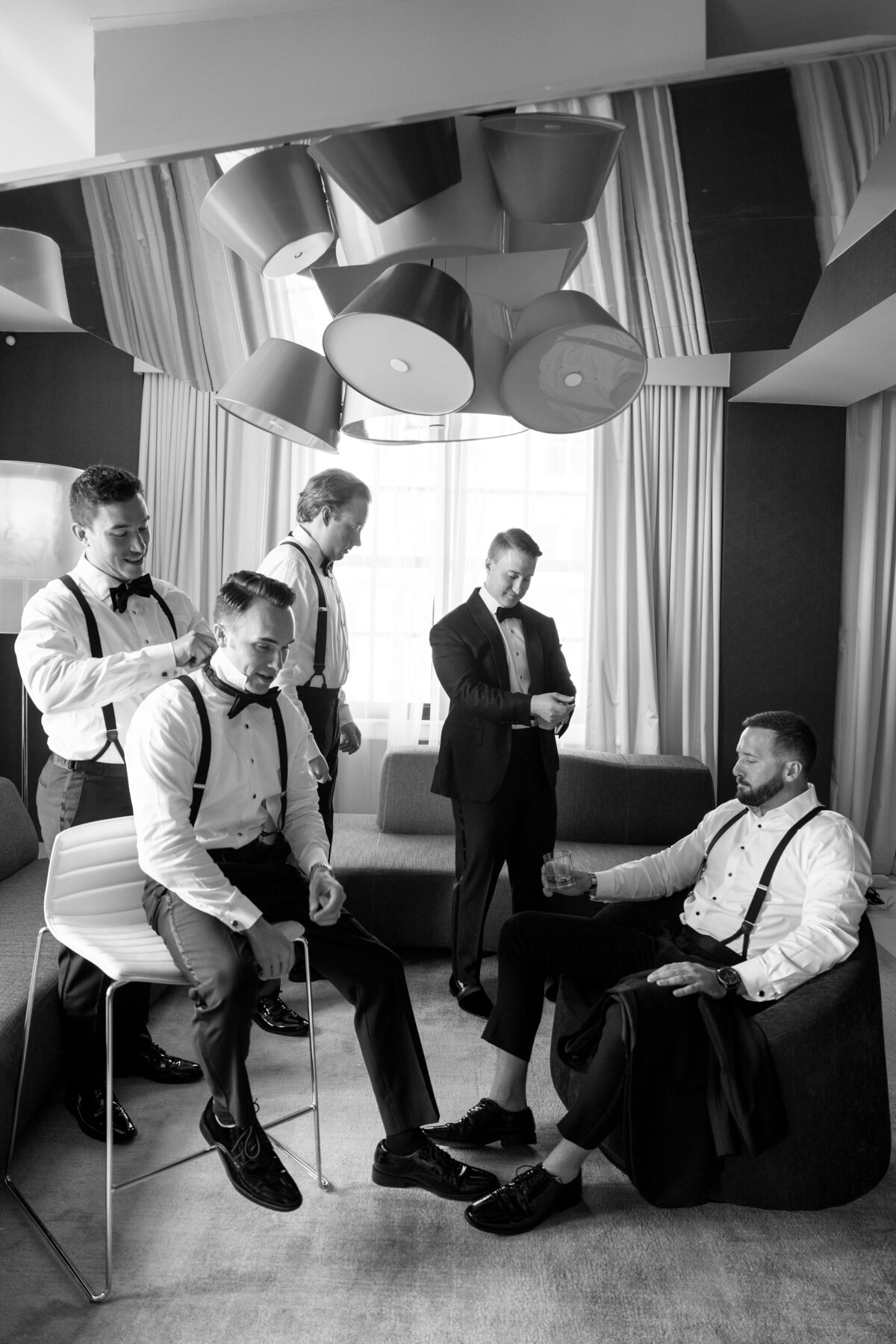A black and white photo of five men in formal attire, including bow ties and suspenders, preparing in a room. One is seated on a stool being assisted with his tie, while others are adjusting their outfits. They appear relaxed and engaged in conversation.