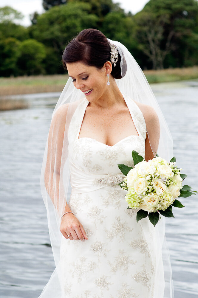 brunette bride wearing a lace, halter neck wedding dress with diamante hairpiece and veil, holding a white flower bouquet beside a lake in Killarney