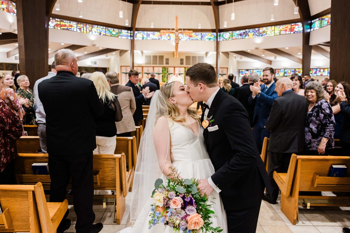 Bride and groom kissing in a church aisle, coordinated by a wedding planner from Des Moines, surrounded by guests clapping, with stained glass windows in the background.