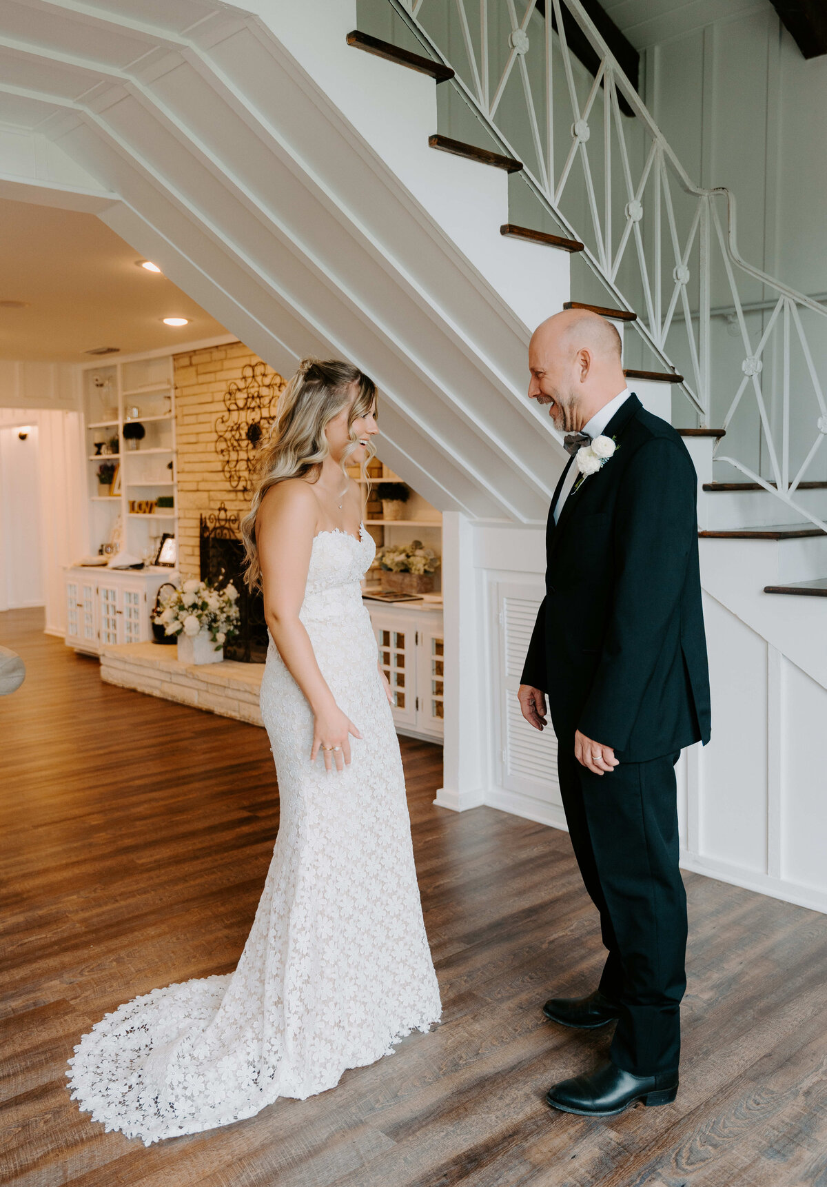 A bride in a lace gown and a smiling man in a black suit with a white rose boutonniere share a warm, emotional moment in a cozy interior near a staircase.
