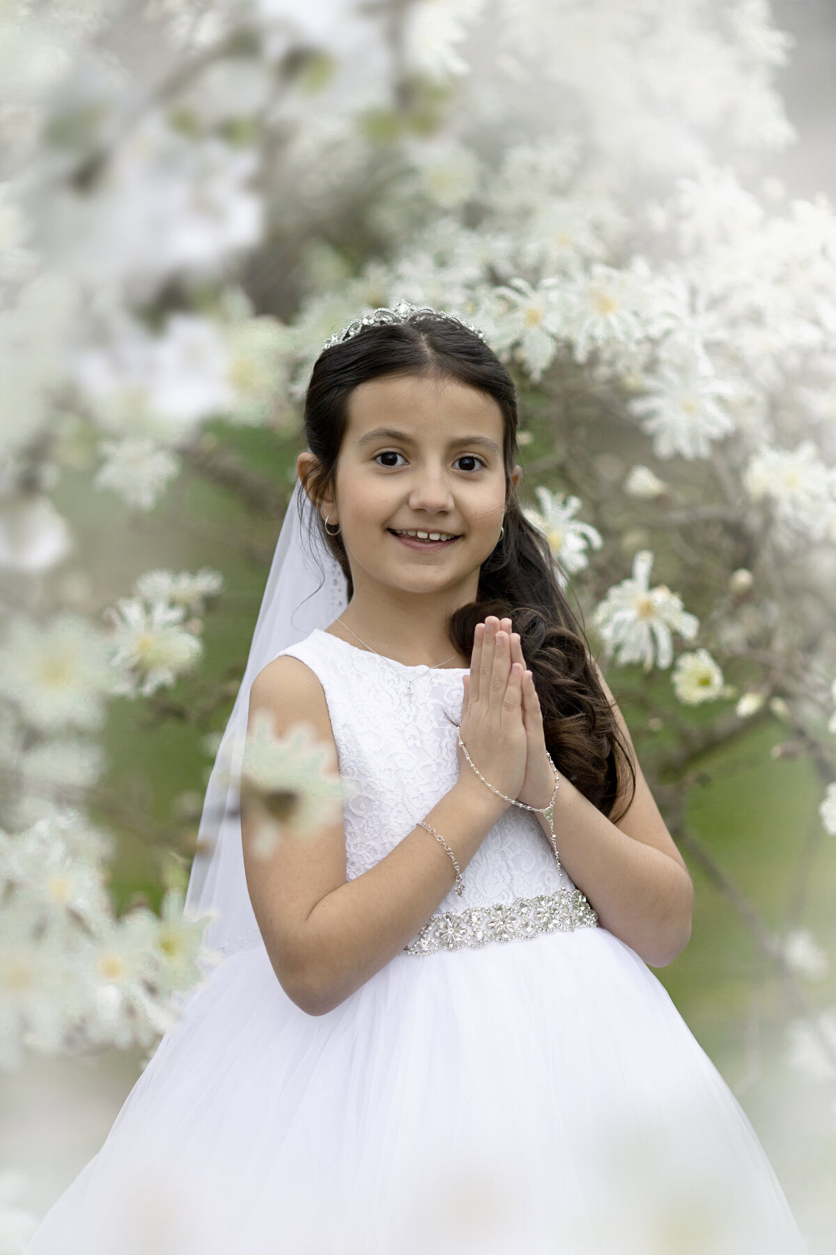 A young girl prays while standing in a white communion dress under a white blooming tree