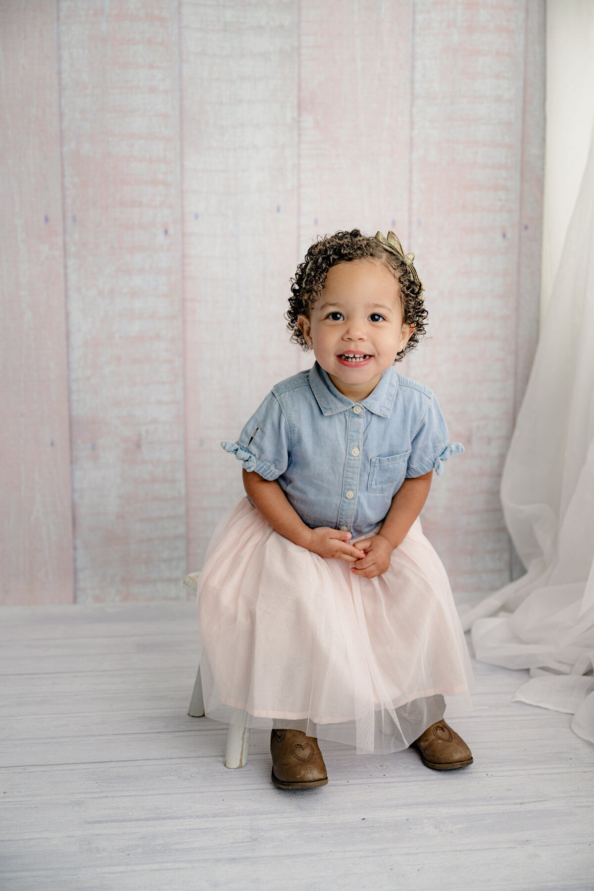 Little girl is dressed in a dress with blue jean top and a skirt bottom on a pink background with sheer white curtains beside her sitting on a bench laughing during her 2-year-old Milestone session