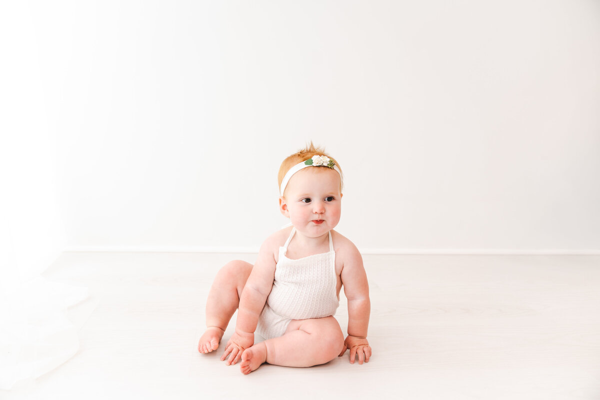 A curious baby sits on the floor, wearing a white onesie and a cute headband, with a soft, neutral background accentuating the child's innocence and charm. Taken by Fig and Olive Photography, Minneapolis Baby Photographer.