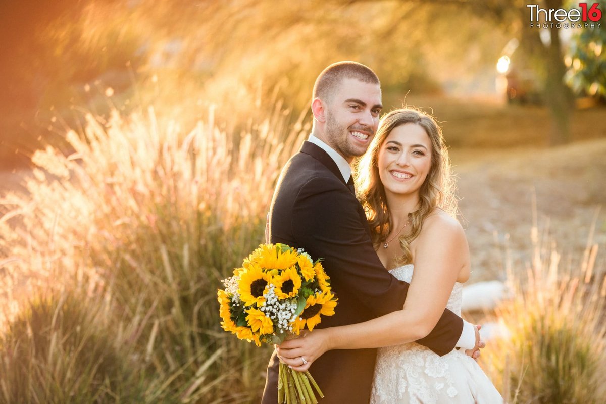 Bride and Groom embrace each other in an open field of brush while she holds her sunflower bouquet