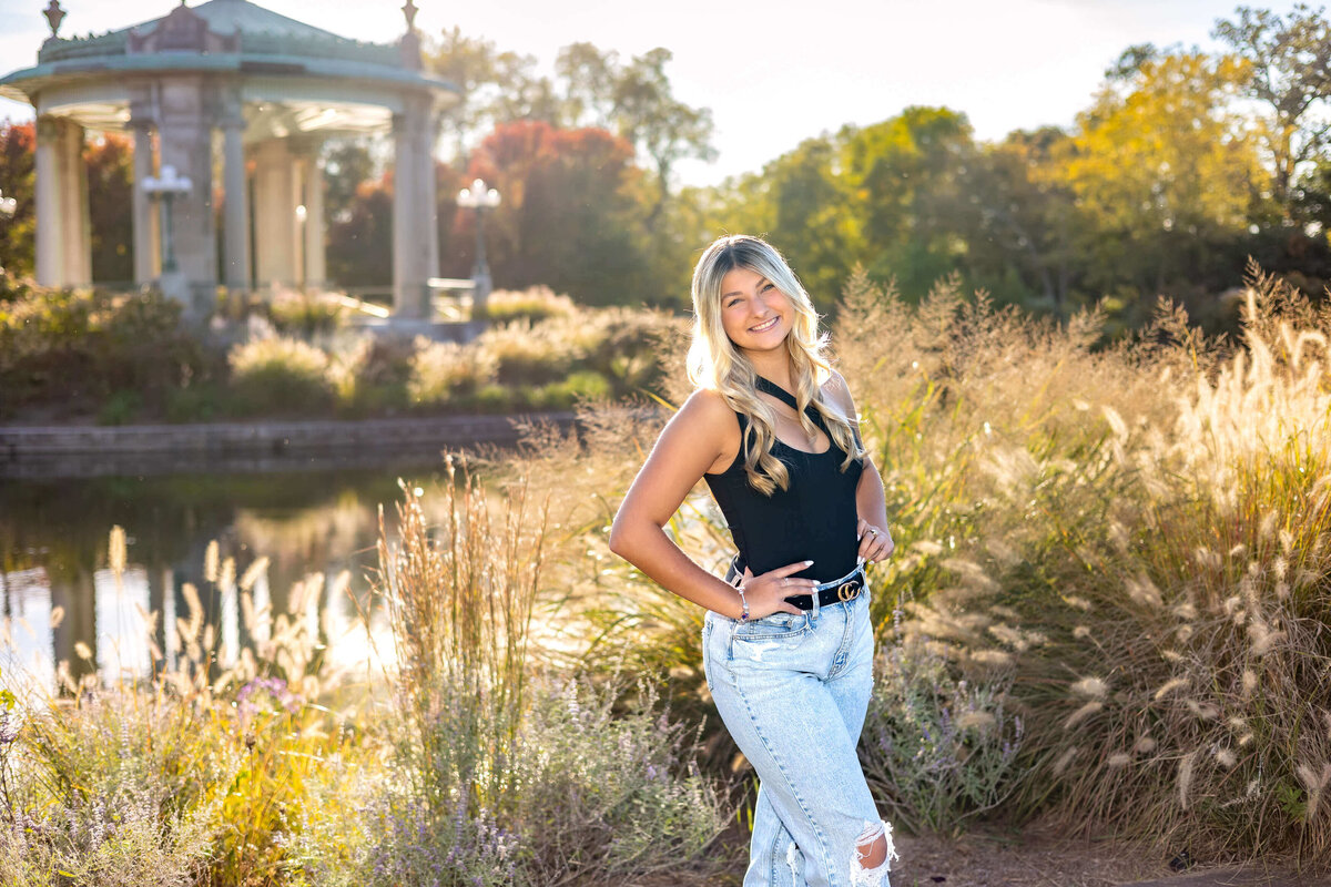 a high school senior girl with long blonde hair standing with her hands on her hips with the sun back lit behind her and lots of tall grass, a gazebo, and  a pond in the background