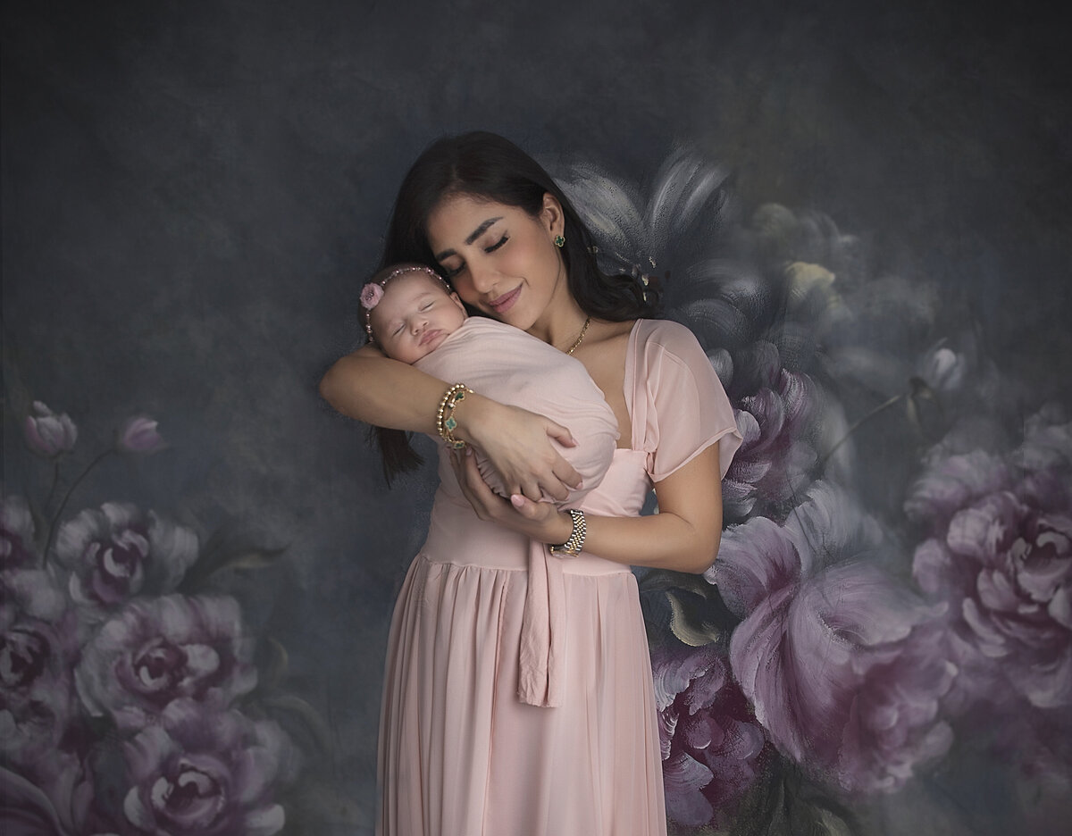 A mom and newborn girl both dressed in pink are posed in front of a floral backdrop posed in a loving embrace in Ottawa.