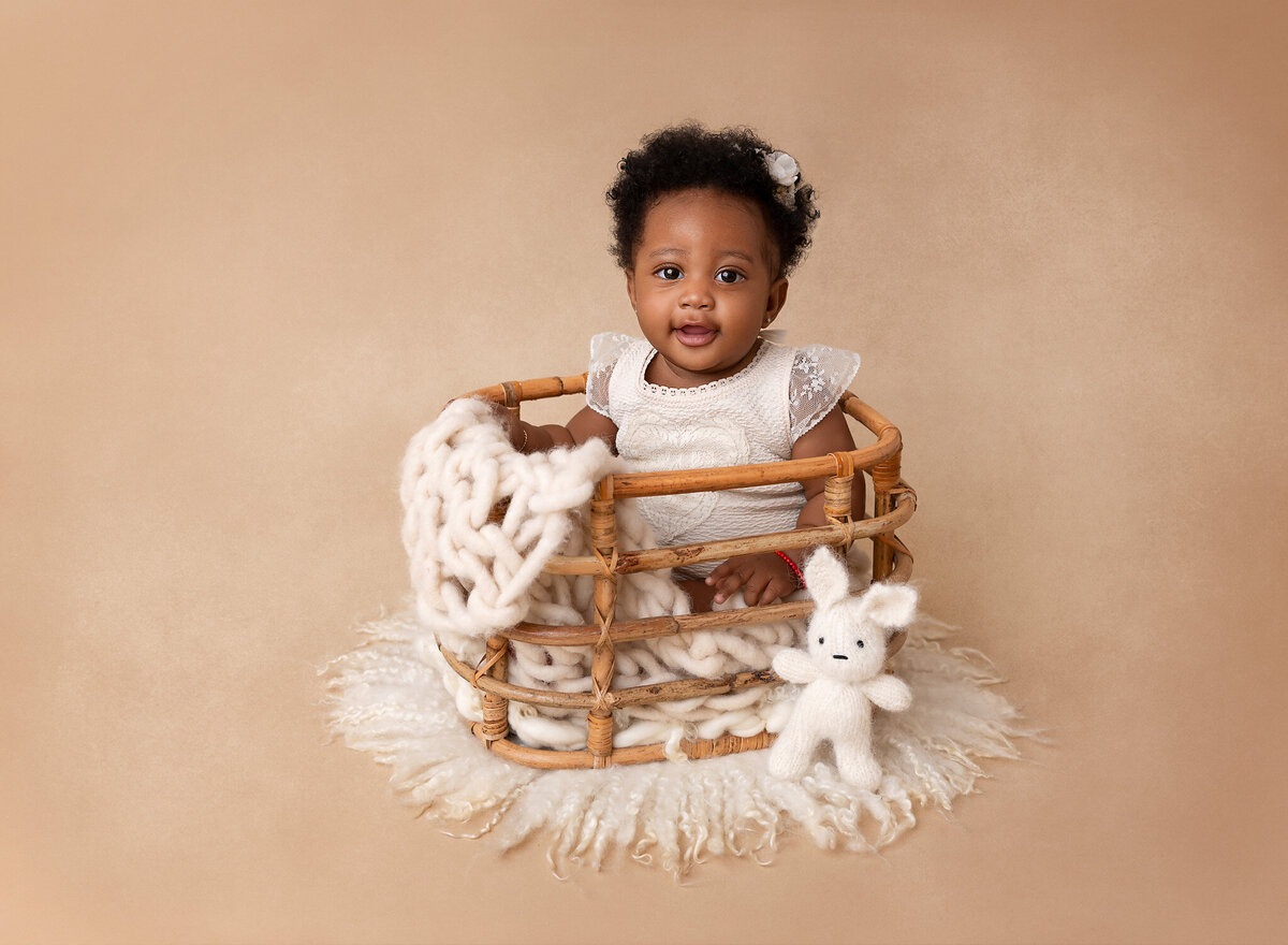Baby girl sitting in a bamboo basket draped with white blankets for a 6-month baby milestone photoshoot. Baby is wearing a white lace romper and smiling at teh camera.