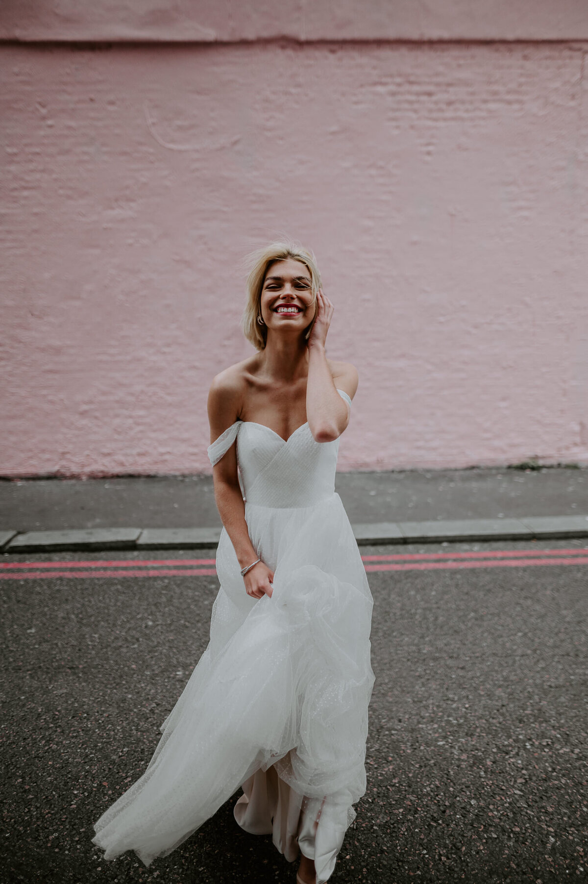 A bride in a shoulder cut Justin Alexander wedding dress tuckers her short hair behind her ear whilst standing in front of a pink walk in Shoreditch, London.