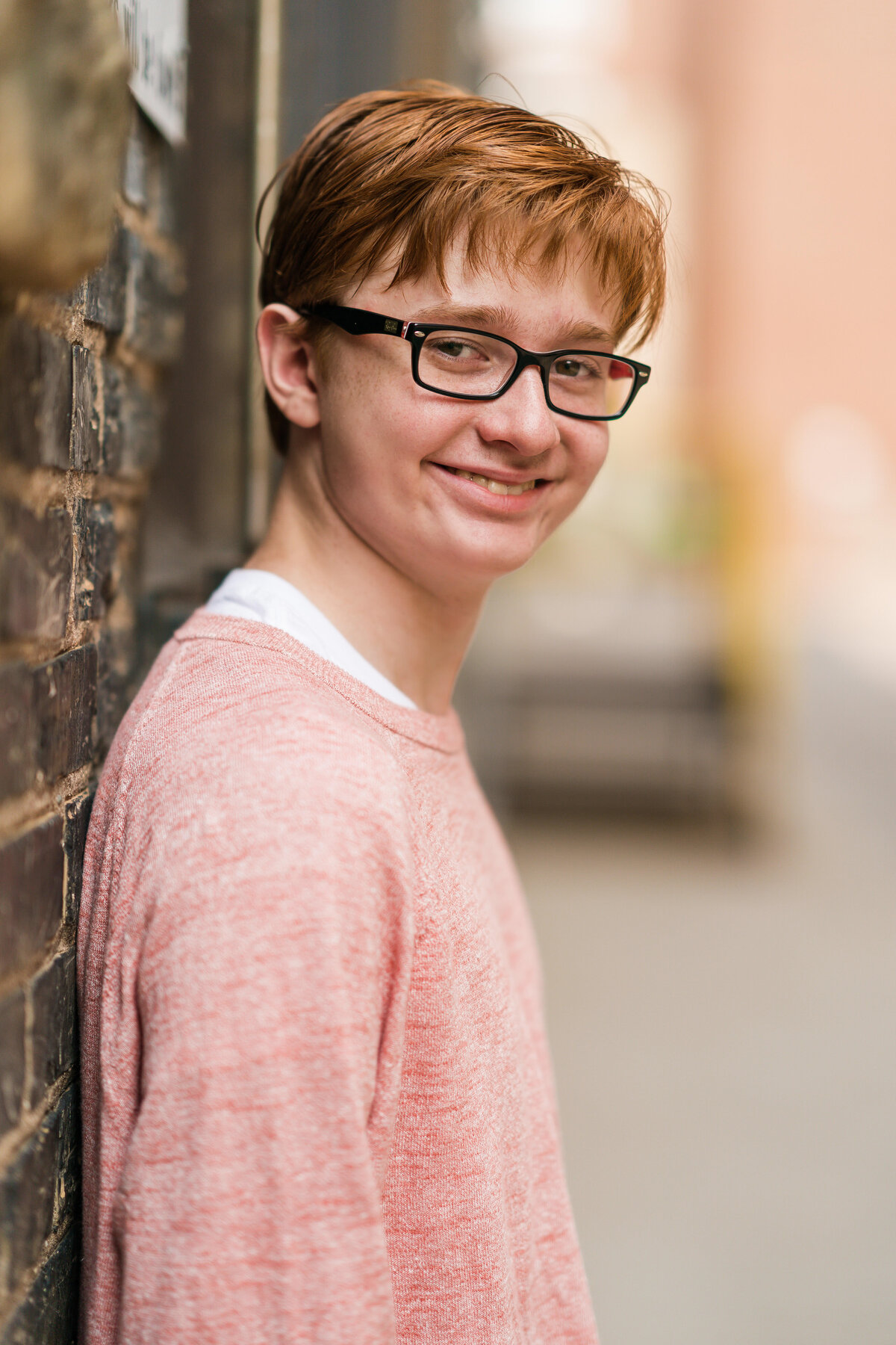 Senior boy with glasses dressed in pink shirt leans against a brick wall.
